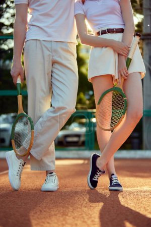 Photo for Cropped image of man and woman in stylish, light, casual clothes posing with tennis racket on open air court during sunset. Concept of casual fashion, sport, active lifestyle, hobby, leisure time, ad - Royalty Free Image
