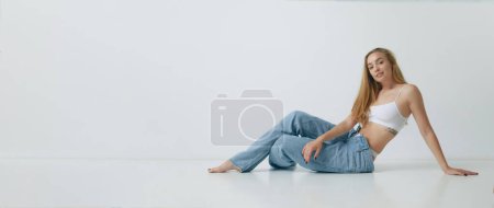 Photo for Beautiful young blonde woman with slim body posing in white top and jeans against grey studio background. Concept of beauty, body and skin care, health, fitness, wellness, ad. Banner - Royalty Free Image