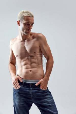 Photo for Portrait of handsome young man with blonde hair, musuclar fit body posing shirtless in jeans against grey studio background. Concept of male natural beauty, body care, health, sport, fashion, ad - Royalty Free Image