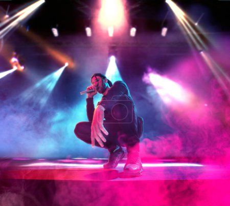 Photo for Artistic young man in stylish clothes with dreads performing on stage at nightclub, singing rap over multicolor neon light. Concept of music, performance, art, talent, nightlife, joy, party, lifestyle - Royalty Free Image