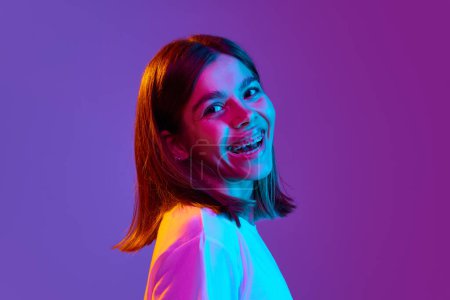 Photo for Close-up portrait of pretty, happy young girl with braces looking at camera and laughing against gradient purple background in neon light. Concept of emotions, youth, feelings, fashion, lifestyle, ad - Royalty Free Image