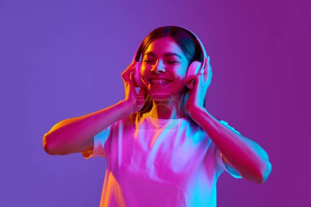 Photo for Portrait of smiling, happy, young girl listening to music in headphones against gradient purple background in neon light. Concept of human emotions, youth, feelings, fashion, lifestyle, ad - Royalty Free Image