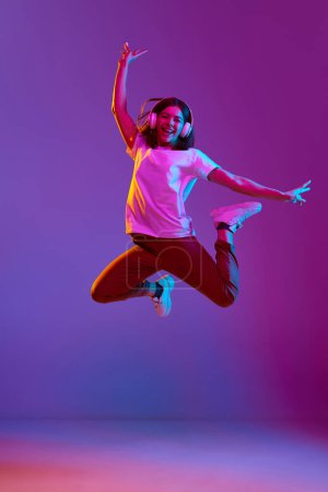 Photo for Full-length portrait of young, happy, smiling girl in casual clothes as headphones jumping over gradient purple background in neon light. Concept of emotions, youth, feelings, fashion, lifestyle, ad - Royalty Free Image