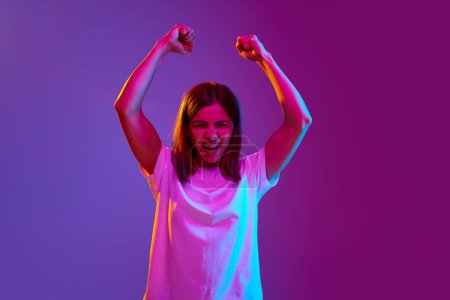 Photo for Motivation and ambitions. Portrait of young girl raising hands up, showing power and strength on gradient purple background in neon light. Concept of emotions, youth, feelings, fashion, lifestyle, ad - Royalty Free Image