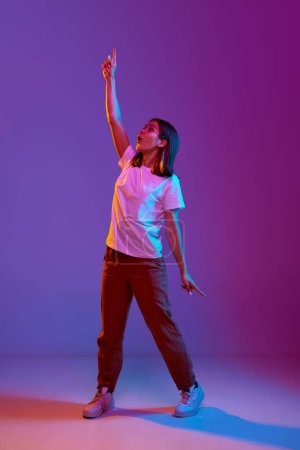 Photo for Full-length portrait of young girl in casual comfortable clothes standing with hand raised over gradient purple background in neon light. Concept of emotions, youth, feelings, fashion, lifestyle, ad - Royalty Free Image