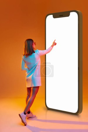 Photo for Online shopping. Young girl in white dress touching screen of 3D model of mobile phone against orange studio background in neon light. Concept of business, emotions, youth, feelings, fashion, ad - Royalty Free Image