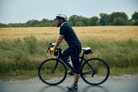 Téléchargez les photos : Jeune homme barbu, cycliste en uniforme, casque et lunettes debout avec vélo sur la route autour du fond du terrain. Concept de sport, hobby, loisir, entraînement, santé, vitesse, endurance, publicité - en image libre de droit