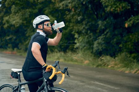 Téléchargez les photos : Jeune homme barbu, cycliste en uniforme, casque et lunettes au bord de la route et eau potable. Rafraîchissements et repos. Concept de sport, hobby, loisir, entraînement, santé, vitesse, endurance - en image libre de droit