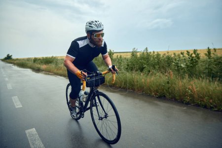 Photo for Bearded young man, cyclist in helmet, glasses and uniform riding bike on wet road in cloudy chill evening. Concept of sport, hobby, leisure activity, training, health, speed, endurance, ad - Royalty Free Image