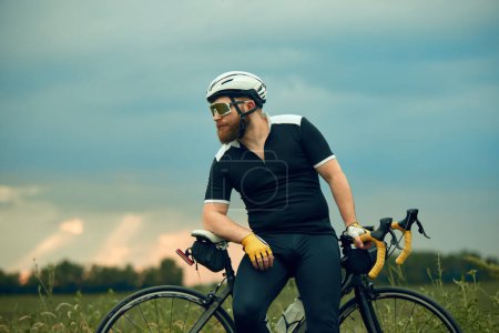 Téléchargez les photos : Homme barbu en vêtements de sport, lunettes et casque, assis à vélo sur la route sur le terrain dans la soirée froide d'été. Concept de sport, hobby, loisir, entraînement, santé, vitesse, endurance, publicité - en image libre de droit
