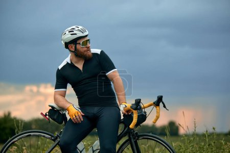 Téléchargez les photos : Élégant, barbu, jeune homme en tenue de sport et casque, assis à vélo sur la route sur le terrain dans la soirée froide d'été. Concept de sport, hobby, loisir, entraînement, santé, vitesse, endurance, publicité - en image libre de droit