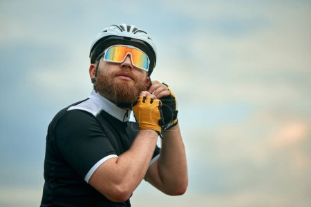 Téléchargez les photos : Jeune homme barbu, cycliste en lunettes de sport dégradées, portant un casque sur fond de ciel nuageux. Concept de sport, hobby, loisir, entraînement, santé, vitesse, endurance, publicité - en image libre de droit