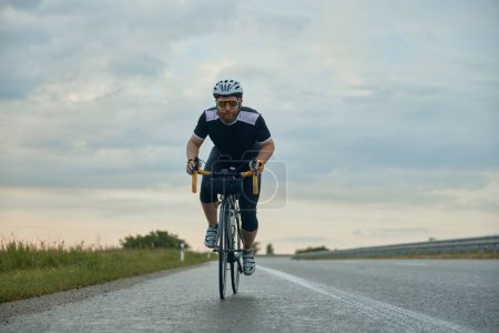 Téléchargez les photos : Image dynamique de jeune homme barbu, cycliste en casque à vélo sur route vide dans la soirée. Week-ends actifs. Concept de sport, hobby, loisir, entraînement, santé, vitesse, endurance, publicité - en image libre de droit