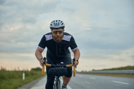 Photo for Dynamic image of young bearded man , cyclist in helmet riding bike on empty road in the evening. Concentration. Concept of sport, hobby, leisure activity, training, health, speed, endurance, ad - Royalty Free Image