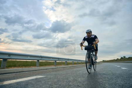 Téléchargez les photos : Image dynamique de jeune homme sportif barbu, cycliste en casque à vélo sur route vide dans la soirée. Concept de sport, hobby, loisir, entraînement, santé, vitesse, endurance, publicité - en image libre de droit
