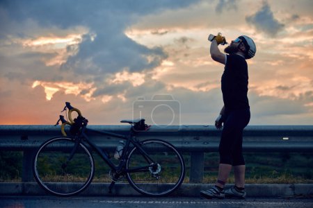 Téléchargez les photos : Jeune homme sportif, cycliste en casque et vêtements confortables debout sur la route avec vélo et eau potable. Coucher de soleil. Concept de sport, hobby, loisir, entraînement, santé, vitesse, endurance - en image libre de droit