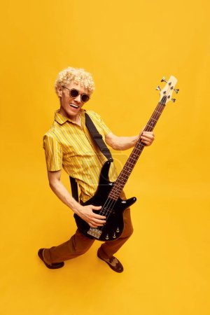 Photo for Positive, talented, young man with curly blonde hair smiling, playing guitar against yellow studio background. Concept of music, talent, hobby, entertainment, festival, performance, ad - Royalty Free Image