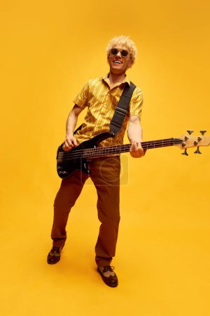 Photo for Full-length image of young smiling blonde man with curly hair in retro clothes playing guitar over yellow studio background. Concept of music, talent, hobby, entertainment, festival, performance, ad - Royalty Free Image