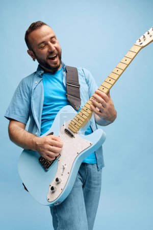 Photo for Smiling bearded man in casual clothes emotionally playing guitar against blue studio background. Lifestyle. Concept of music, talent, hobby, entertainment, festival, performance, ad - Royalty Free Image
