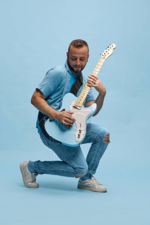 Photo for Young, smiling happy man in casual clothes emotionally, cheerfully playing guitar against blue studio background. Concept of music, talent, hobby, entertainment, festival, performance, ad - Royalty Free Image