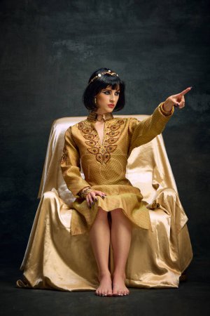 Foto de Retrato de una hermosa mujer joven en imagen de Cleopatra, reina en elegante vestido de oro sentado y al mando sobre fondo oscuro vintage. Concepto de cultura antigua, historia, comparación de épocas, arte - Imagen libre de derechos