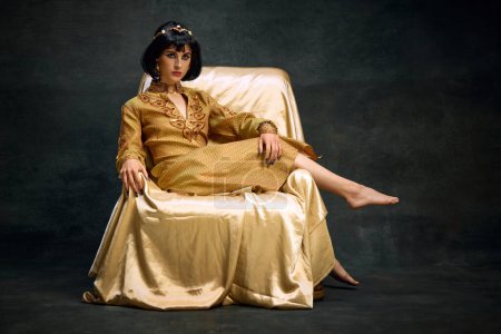 Photo for Portrait of elegant, beautiful woman in image of antique queen, Cleopatra in olden clothes sitting on armchair on dark vintage background. Concept of antique culture, history, comparison of eras, art - Royalty Free Image