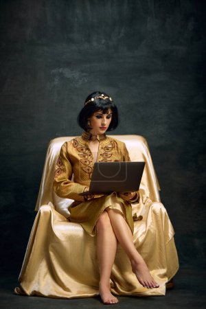 Téléchargez les photos : Portrait de belle jeune femme à l'image de la reine, Cléopâtre assise en robe dorée et travaillant sur ordinateur portable sur fond vintage sombre. Concept de culture antique, histoire, comparaison des époques, art - en image libre de droit