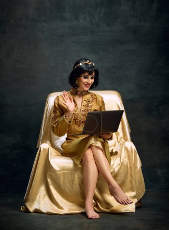 Photo for Portrait of beautiful young woman in image of queen, Cleopatra sitting and talking on vide via laptop against dark vintage background. Concept of antique culture, history, comparison of eras, art - Royalty Free Image