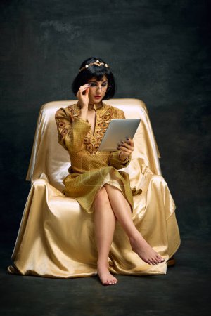 Foto de Joven mujer hermosa, reina, Cleopatra en vestido de oro sentado con la tableta y trabajando sobre fondo oscuro vintage. Proyectos en línea. Concepto de cultura antigua, historia, comparación de épocas, arte - Imagen libre de derechos