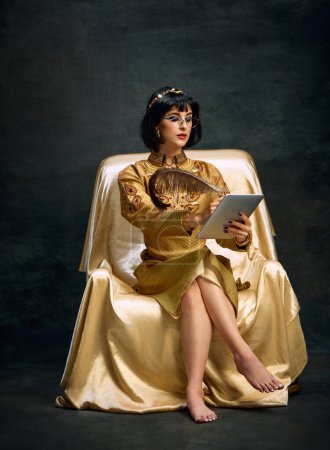 Photo for Portrait of young beautiful woman, queen, Cleopatra in golden dress sitting with tablet and writing letter against dark vintage background. Concept of antique culture, history, comparison of eras, art - Royalty Free Image