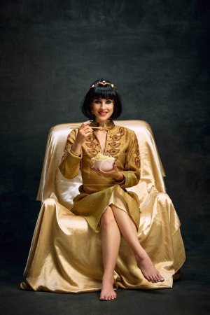 Foto de Retrato de una joven sonriente en imagen de la reina egipcia, Cleopatra comiendo fideos instantáneos sobre fondo oscuro vintage. Concepto de cultura antigua, historia, comparación de épocas, arte, belleza, ad - Imagen libre de derechos