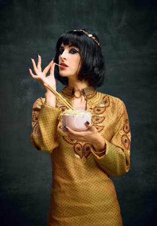 Téléchargez les photos : Portrait de belle jeune femme à l'image de la reine égyptienne, Cléopâtre mangeant des nouilles instantanées sur fond sombre vintage. Concept de culture antique, histoire, comparaison des époques, art, beauté, annonce - en image libre de droit