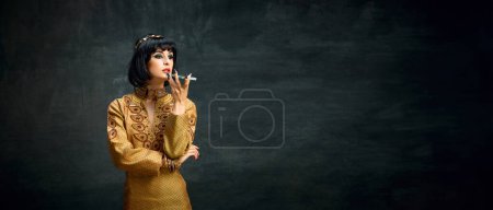 Photo for Portrait of young woman with expressive makeup in image of Cleopatra talking on mobile phone against dark vintage background. Concept of antique culture, history, comparison of eras, art, beauty, ad - Royalty Free Image