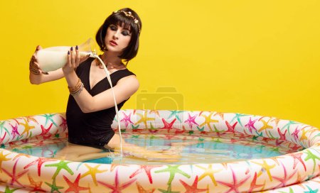 Photo for Young artistic woman in image of queen, Cleopatra in swimming pool and pouring milk inside against yellow studio background. Concept of antique culture, history, comparison of eras, art, beauty, ad - Royalty Free Image