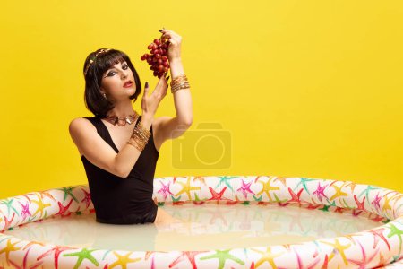 Foto de Hermosa joven en imagen de Cleopatra sentada en la piscina con leche y comiendo uvas sobre fondo amarillo del estudio. Concepto de cultura antigua, historia, comparación de épocas, arte, belleza - Imagen libre de derechos