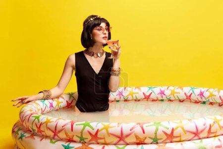 Photo for Beautiful young woman in image of Cleopatra sitting in swimming pool with milk and drinking cocktail against yellow studio background. Concept of antique culture, history, comparison of eras, art, ad - Royalty Free Image