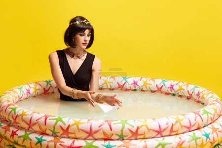 Foto de Mujer joven y bonita en la imagen de Cleopatra sentada en la piscina con leche y jugando con un barco de papel sobre fondo amarillo. Sueños. Concepto de cultura antigua, historia, comparación de épocas, arte - Imagen libre de derechos