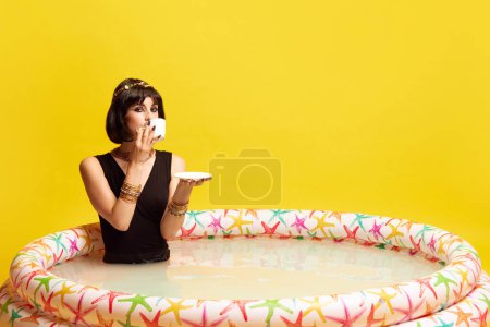 Téléchargez les photos : Jolie jeune femme à l'image de Cléopâtre assise dans la piscine avec du lait et buvant du café sur fond jaune studio. Concept de culture antique, histoire, comparaison des époques, art, beauté, annonce - en image libre de droit
