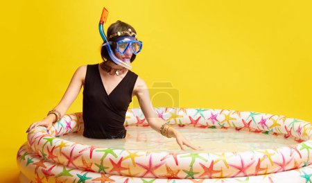 Photo for Beautiful young woman in image of Cleopatra sitting in swimming pool with diving equipment against yellow studio background. Vacation. Concept of antique culture, history, comparison of eras, art, ad - Royalty Free Image