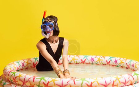 Foto de Hermosa joven en imagen de Cleopatra sentada en la piscina con equipo de buceo sobre fondo amarillo estudio. Vacaciones. Concepto de cultura antigua, historia, comparación de épocas, arte, ad - Imagen libre de derechos