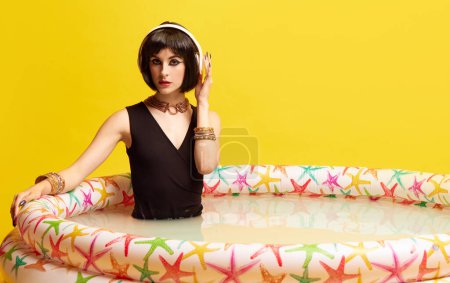 Foto de Hermosa joven en imagen de Cleopatra sentada en la piscina y escuchando música en auriculares sobre fondo amarillo del estudio. Concepto de cultura antigua, historia, comparación de épocas, arte - Imagen libre de derechos