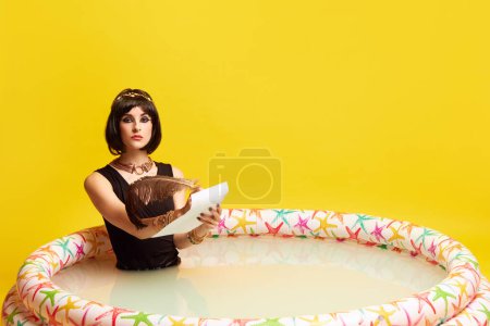 Téléchargez les photos : Jeune femme sérieuse à l'image de Cléopâtre assise dans la piscine et écrivant une lettre sur fond de studio jaune. Concept de culture antique, histoire, comparaison des époques, art, beauté, annonce - en image libre de droit