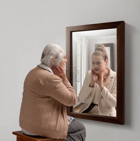Photo for Creative conceptual college. Senior woman looking in mirror with reflection of her young self. Aging process. Concept of present, past and future, age, lifestyle, memories, generation, ad - Royalty Free Image