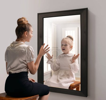 Kreative konzeptionelle Collage. Aufgeregtes junges Mädchen, das in den Spiegel schaut und das Spiegelbild des kleinen Mädchens, ihres kindlichen Selbst, sieht. Konzept der Gegenwart, Vergangenheit und Zukunft, Alter, Lebensstil, Erinnerungen, Generation, Anzeige