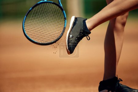 Foto de Imagen recortada de piernas femeninas en zapatillas deportivas y raqueta de tenis. Cayendo por la arena. Oferta de entrenamiento en pista de tenis al aire libre. Concepto de deporte, hobby, estilo de vida activo, salud, resistencia y fuerza, ad - Imagen libre de derechos