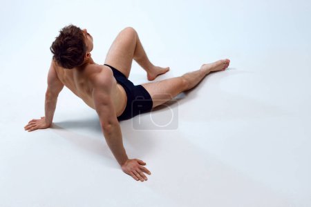 Téléchargez les photos : Jeune homme au corps athlétique, musclé et en forme couché sur le sol, posant torse nu en sous-vêtements sur fond de studio blanc. Concept d'hommes beauté, mode de vie sportif et sain, corps athlétique - en image libre de droit