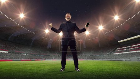 Photo for Happy, lucky, excited businessman standing with mobile phone, feeling success and win of money on sports betting. 3D arena background. Concept of sport, fan, betting, finances, gambling, bookmaker - Royalty Free Image