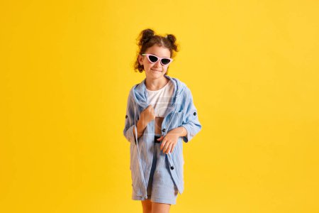Téléchargez les photos : Portrait de belle petite fille, enfant aux cheveux bouclés, en lunettes de soleil posant sur fond de studio jaune. Style d'été. Concept d'émotions, enfance, éducation, mode, mode de vie, publicité - en image libre de droit