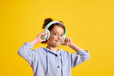 Foto de Retrato de niña positiva escuchando música en auriculares y sonriendo sobre fondo amarillo del estudio. Concepto de emociones, infancia, educación, moda, estilo de vida, anuncio - Imagen libre de derechos