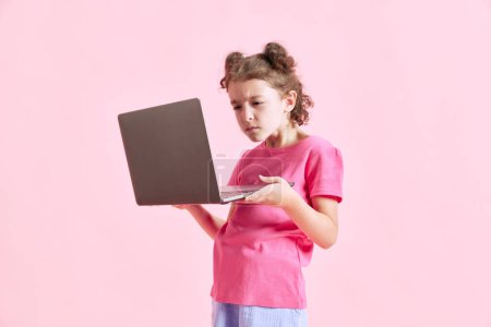 Photo for Portrait of child, little girl with concentrated face looking on laptop against pink studio background. Online lessons. Concept of emotions, childhood, education, fashion, lifestyle, ad - Royalty Free Image